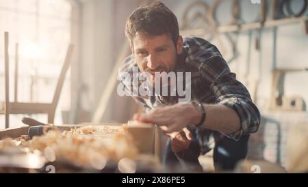 Close Up Portrait of a Handsome Young Artisan Craftsman in Checkered Shirt Using Hand Plane to Shape a Wood Bar. Carpenter Working on a Project in a Loft Studio with Tools on Walls. Stock Photo