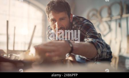 Handsome Young Artisan Craftsman in Checkered Shirt Using Hand Plane to Shape a Wood Bar. Carpenter Working on a Project in a Loft Studio with Tools on Walls. Stock Photo