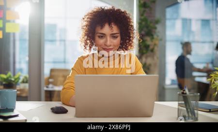 Beautiful Middle Eastern Manager Sitting at a Desk in Creative Office. Young Stylish Female with Curly Hair Using Laptop Computer in Marketing Agency. Colleagues Working in the Background Stock Photo