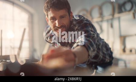 Young Artisan Craftsman in Checkered Shirt Using Hand Plane to Shape a Wood Bar. Handsome Carpenter Working on a Project in a Woodworking Workshop. Stock Photo