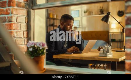 Portrait of a Young Handsome Black Man Sitting at a Desk, Working on Laptop Computer. Freelance Journalist Writing an Article Remotely from Home Office. Stylish Apartment Shot from Window. Stock Photo