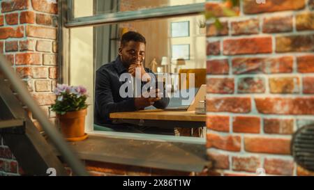 Happy Young Black Male Sitting at Home, Connecting with Friends on Social Media, Using Online Apps on Smartphone. Freelancer Working at Home from His Stylish Living Room. View from Outside the Window. Stock Photo