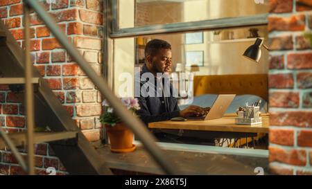Tired African Male Using Laptop at Home. Doing Remote Work During the Day. Frustrated Man Sitting at a Table, Working Long Hours on Computer, Brainstorming Creative Project. Outside the Window Shot. Stock Photo