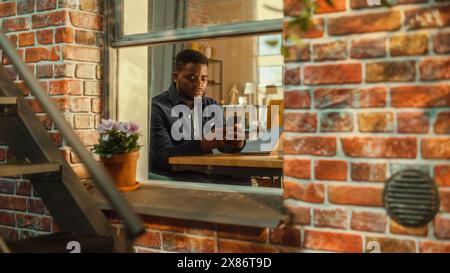 Happy Young Black Male Sitting at Home, Connecting with Friends on Social Media, Using Online Apps on Smartphone. Freelancer Working at Home from His Stylish Living Room. View from Outside the Window. Stock Photo