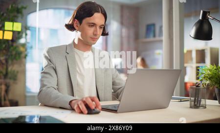 Creative Project Manager Sitting at a Desk, Working on a Laptop Computer in an Office. Handsome Marketing Specialist in a Casual Jacket Planning a Project, Browsing Internet and Writing Emails. Stock Photo