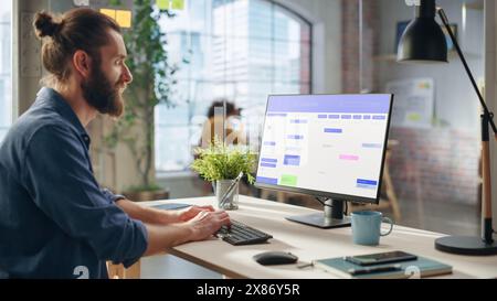 Handsome Long-Haired Bearded Manager Working at a Desk in Creative Office, Using Desktop Computer with a Calendar Software on Screen. Colleagues Working in the Background in Marketing Agency. Stock Photo