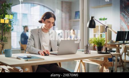 Happy Long-Haired Manager Sitting at a Desk in Creative Office. Young Stylish Man Using Laptop Computer in Marketing Agency. Colleagues Working in the Background. Stock Photo