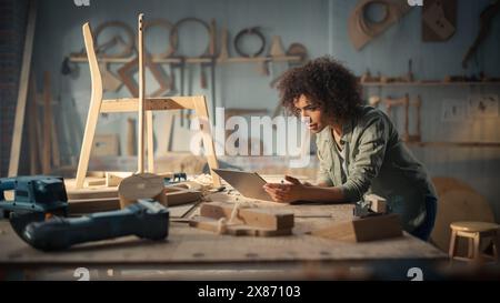 Multiethnic Female Carpenter Using Laptop Computer Brainstorming Creative Ideas Online. Black Female Furniture Designer Working in a Studio in Loft Space with Tools on the Walls. Stock Photo