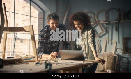 Happy Young Caucasian Man and Multiethnic Woman Using Laptop Computer in Designer Furniture Studio. Carpenters Have Fun, DIscussing and Talking About Upcoming Orders. Stock Photo