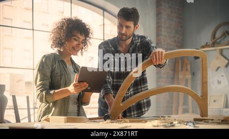 Excited Young Man and Woman Using Tablet Computer, Discussing a Successful Project in a Carpentry Studio. Furniture Designers Working in Loft Studio. Stock Photo