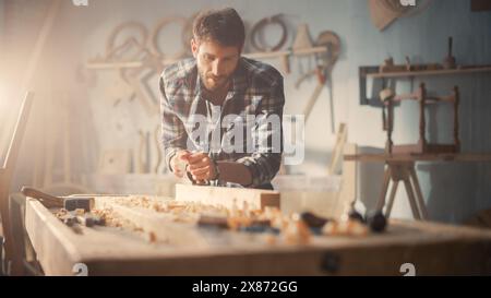 Young Artisan Craftsman in Checkered Shirt Using Hand Plane to Shape a Wood Bar. Handsome Carpenter Working on a Project in a Woodworking Workshop. Stock Photo