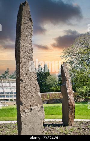 Two tall, weathered stone monoliths standing upright in a grassy area with trees and a building in the background. The sky is colorful with a sunset o Stock Photo