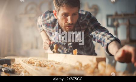 Handsome Young Artisan Craftsman in Checkered Shirt Using Hand Plane to Shape a Wood Bar. Carpenter Working on a Project in a Loft Studio with Tools on Walls. Stock Photo