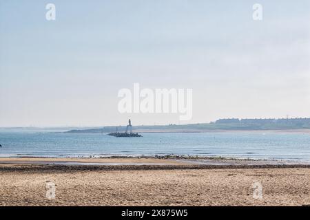 A serene beach scene with a lighthouse on a small rocky island in the distance. The sandy beach is in the foreground, and calm blue waters stretch out Stock Photo