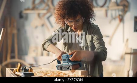 Multiethnic Woman Carpenter Wearing Protective Safety Glasses and Using Electric Belt Sander to Grind a Wood Block. Artist or Furniture Designer Working on a Product Idea in a Workshop. Stock Photo