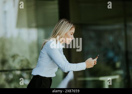 A focused woman in a white blouse engrossed in her smartphone outside a sleek modern office building on a sunny afternoon. Stock Photo
