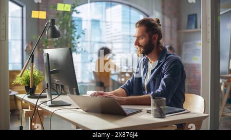 Stylish Long-Haired Bearded Specialist Sitting at a Desk in Creative Agency. Young Stylish Man Working on a Laptop Computer Connected to a Second Display. Office Team Members in the Background. Stock Photo