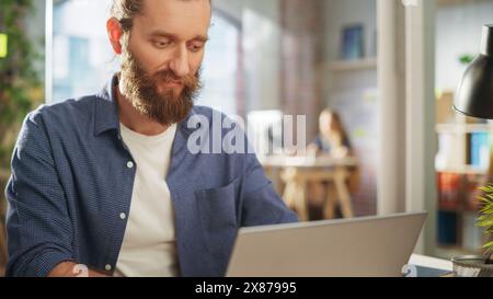 Close Up Portrait of a Handsome Long-Haired Bearded Manager Sitting at a Desk in Creative Office. Stylish Man Using Laptop Computer in Marketing Company. Writing Inspiring Startup Business Ideas. Stock Photo