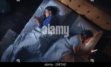 Top View Apartment Bedroom: Caucasian Couple Lying in Bed, Both Using Smartphones. Family of Two Using Mobile Phones to Browse Social Media, Communicate, Search internet, Online Shopping Before Sleep Stock Photo