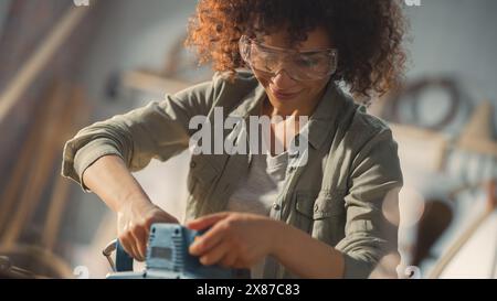 Female Carpenter Wearing Protective Safety Glasses and Using Electric Belt Sander to Work on a Wood Bar. Artist or Furniture Designer Working on a Product Idea in a Workshop. Stock Photo