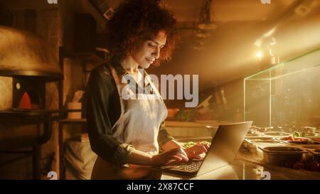 In Restaurant: Beautiful Female Chef Using Laptop Computer. Authentic Pizzeria With Delicious Organic Eco Food. Bi-racial Female Entrepreneur Working in Her Small Business Family Shop Stock Photo