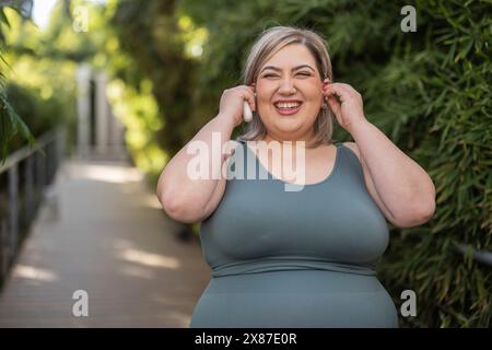 Cheerful curvy woman wearing wireless in-ear headphones near plants Stock Photo