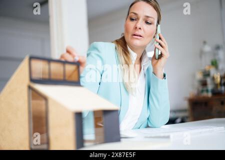 Confident mature architect talking on mobile phone sitting with model house at desk in office Stock Photo