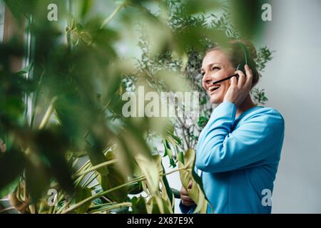 Happy mature businesswoman talking through headset by plants in office Stock Photo