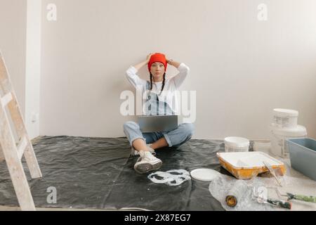 Shocked woman using laptop sitting on plastic near paint equipment in new home Stock Photo