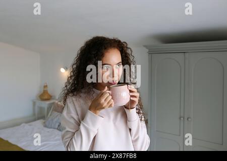Thoughtful young woman drinking coffee in bedroom Stock Photo