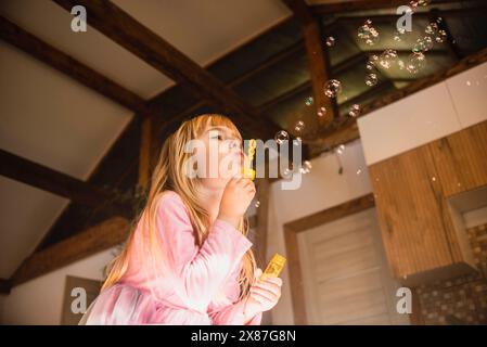Blond girl blowing soap bubbles through wand in kitchen at home Stock Photo