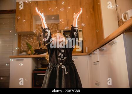 Happy girl smiling and playing with soap bubbles in kitchen at home Stock Photo