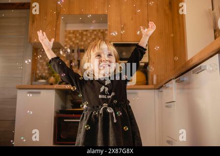 Smiling girl with arms raised playing with bubbles in kitchen at home Stock Photo