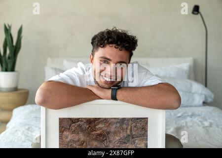 Happy young man sitting with picture frame on bed at home Stock Photo