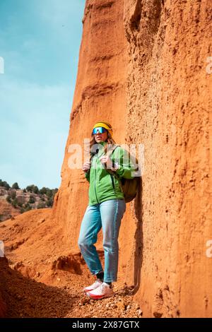 Mature woman standing with backpack by red rocky cliff at canyon on sunny day Stock Photo