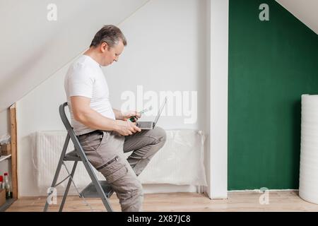 Man holding color swatch and using laptop sitting on stepladder at home Stock Photo