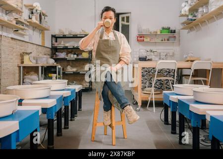Craftsperson drinking coffee sitting on stool amidst pottery wheels at workplace Stock Photo