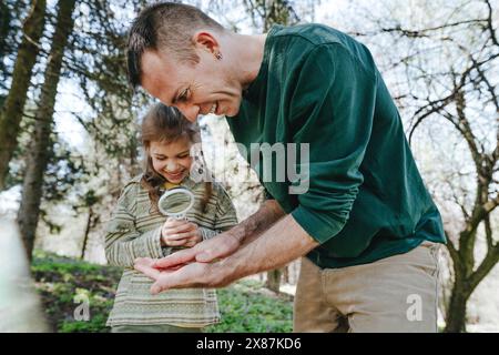 Happy girl examining bug on father's hand with magnifying glass in forest Stock Photo