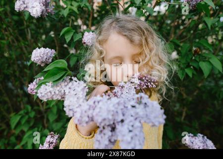 Blond curly haired girl smelling lilacs near plants Stock Photo