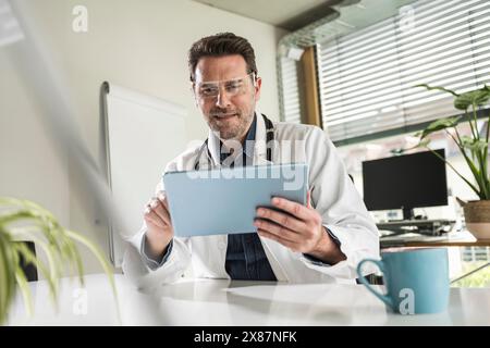 Smiling doctor wearing smart glasses using tablet PC sitting at desk in office Stock Photo