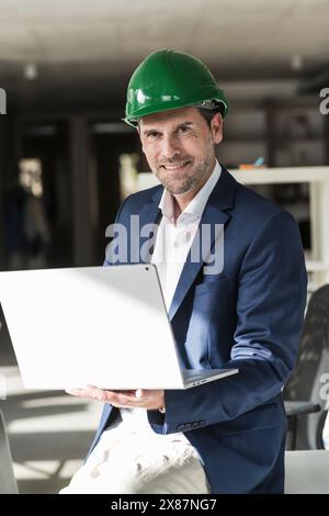Mature engineer wearing hardhat using laptop sitting on desk in office Stock Photo