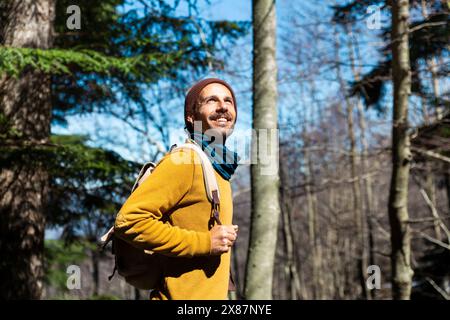 Smiling man with backpack standing in forest at sunny day Stock Photo