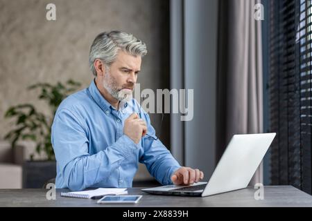 A mature professional is concentrating on his work in an office, using a laptop and looking pensive and focused Stock Photo