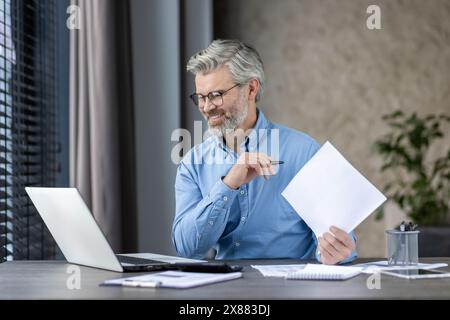 Smiling middle-aged man working at laptop home office hat home office holding papers and wearing glasses. Stock Photo