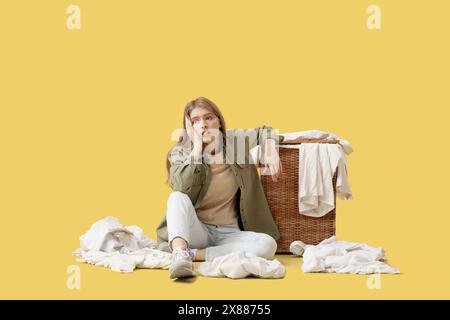 Tired young woman sitting near laundry basket on yellow background Stock Photo