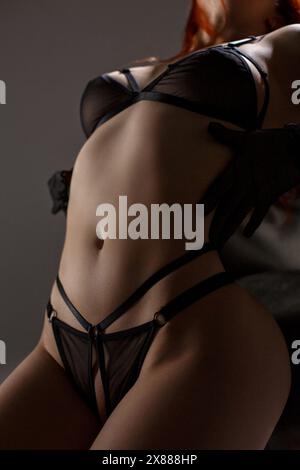 Close up of a young sexy redhead woman in see-through black lingerie  leaning on a chair in studio lights and attractively posing with hands in  black g Stock Photo