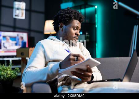 Entrepreneur working from RGB lit home, writing startup ideas on paper while watching video on laptop. BIPOC man feeling delighted after coming up with entrepreneurship plan, using pen to note it down Stock Photo