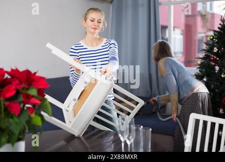 Young woman dusts chair while an elderly mother vacuums room before Christmas Stock Photo