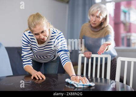 Elderly mother is unhappy with the way her adult daughter dusts the table in room Stock Photo