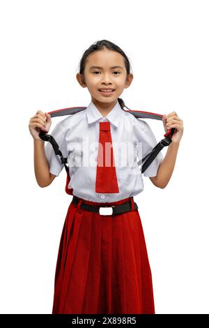 Cheerful young girl stands proudly in her red and white school uniform, playfully pulling on her suspenders, isolated on a white background with a bri Stock Photo